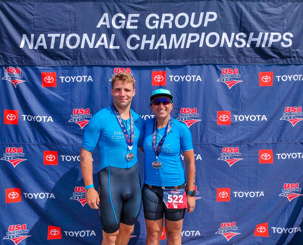 male and female triathletes standing in front of age group national championships sign